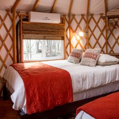 Twin bed with white sheets and a red blanket next to the window of a yurt.