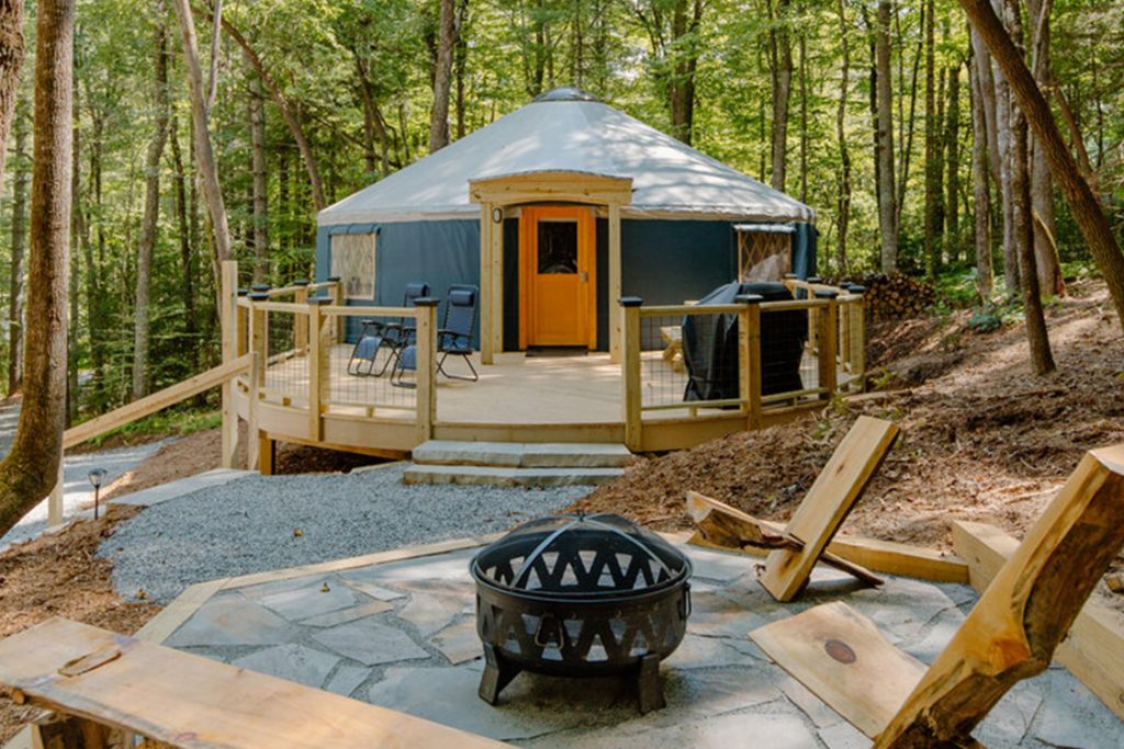 Campfire and log benches in front of a blue Dupont Yurt with a wooden patio.