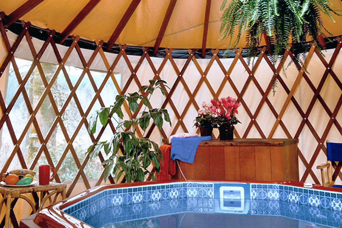 Hot tub inside of a yurt next to a wooden dresser with a blue towel and flowers on top.