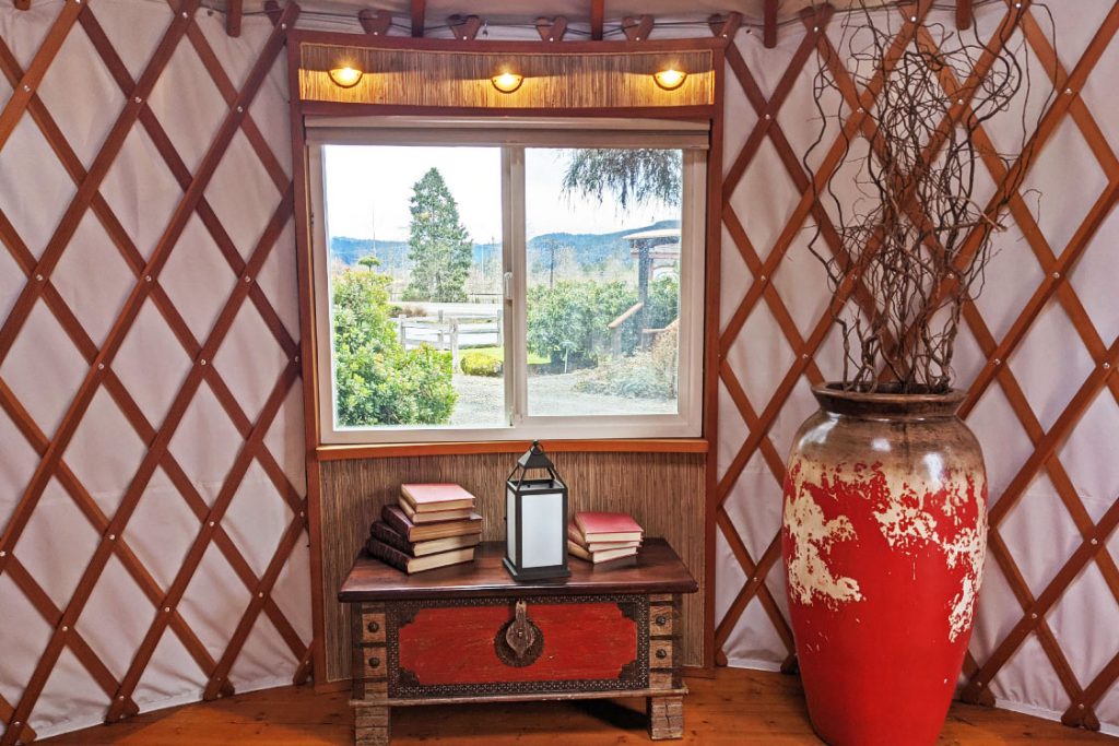 Small table with books and a lamp under a yurt window with a red vase to the right.