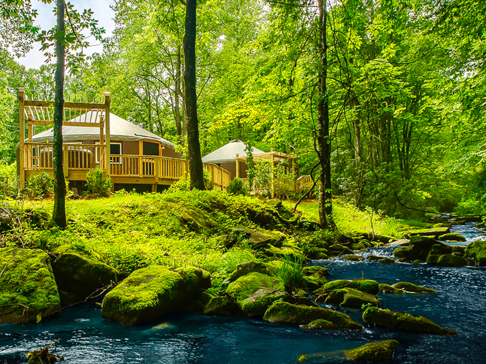 Two Pacific Yurts with wooden decks around the exterior that overlook a river in a forest.