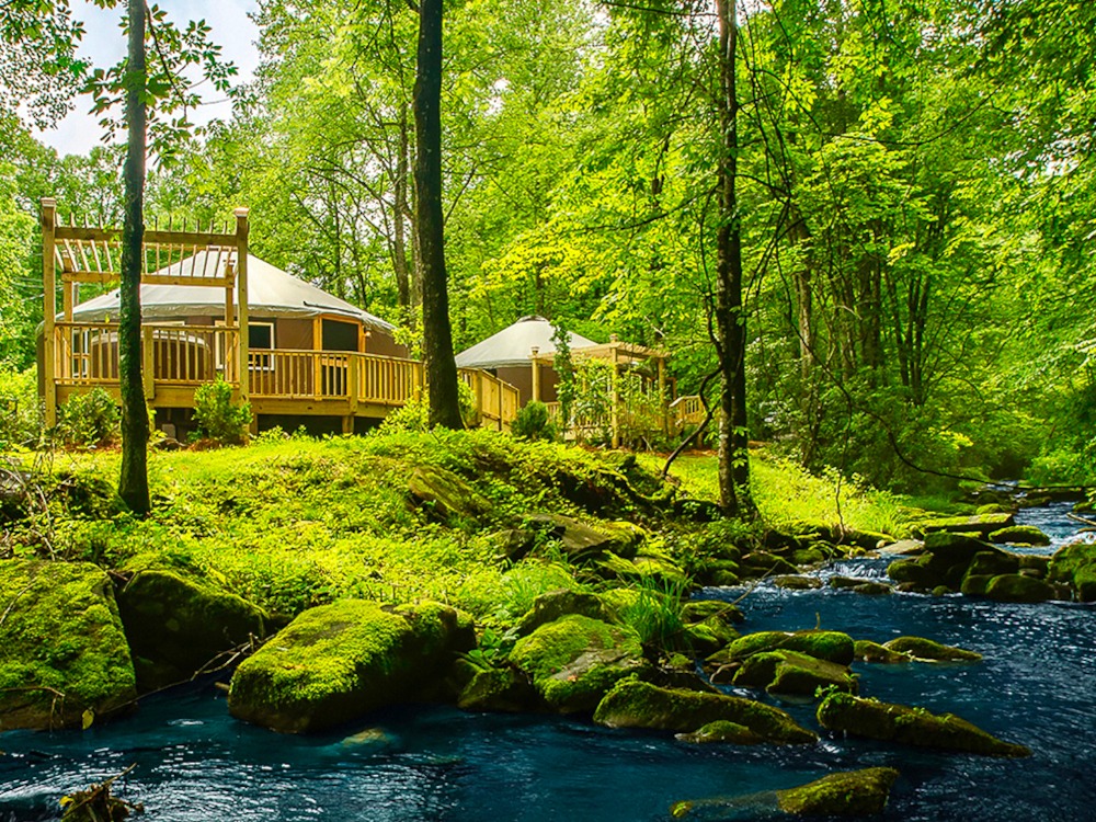 Two Pacific Yurts with wooden decks around the exterior that overlook a river in a forest.