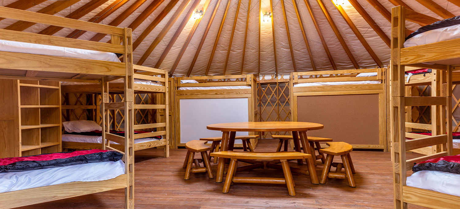 A round wooden table with wooden benches in the middle of a Pacific Yurt living space, surrounded by six bunk beds.
